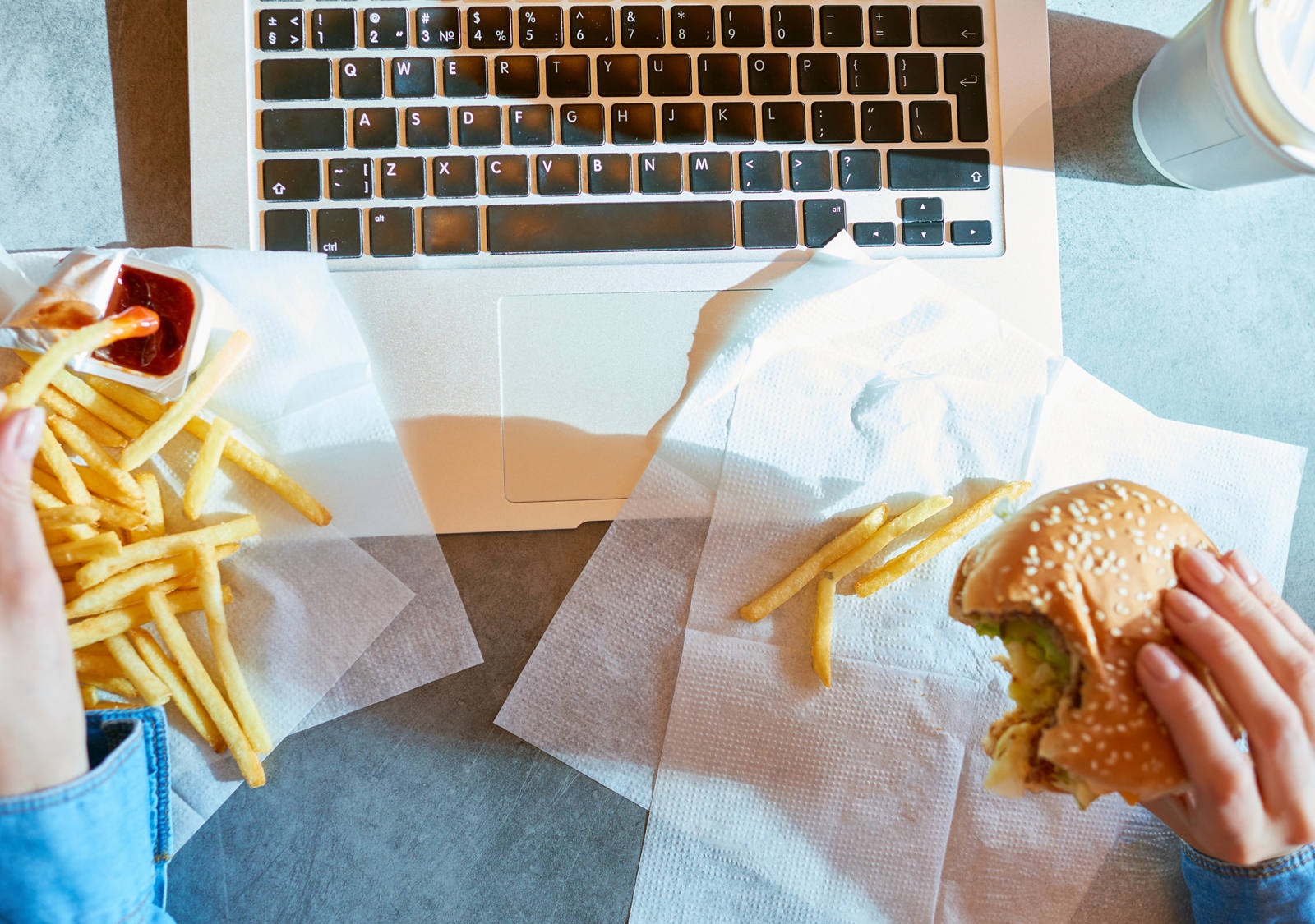 Jemand sitzt am Tisch vor dem Laptop und isst in Hektik Pommes mit Ketchup und einen Burger.