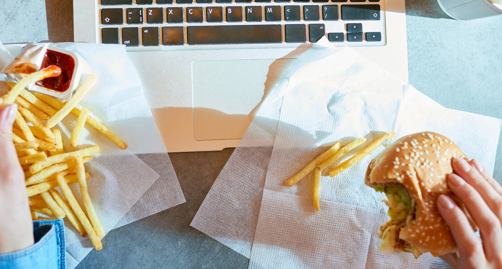 Jemand sitzt am Tisch vor dem Laptop und isst in Hektik Pommes mit Ketchup und einen Burger.
