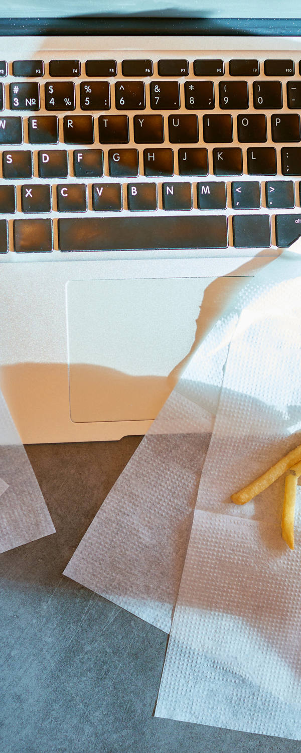 Jemand sitzt am Tisch vor dem Laptop und isst in Hektik Pommes mit Ketchup und einen Burger.