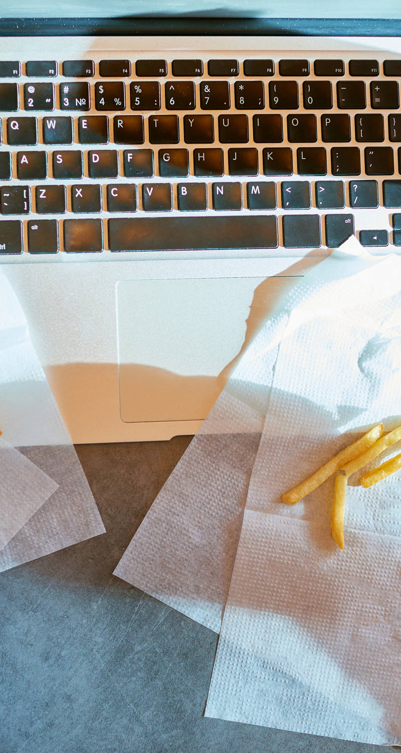 Jemand sitzt am Tisch vor dem Laptop und isst in Hektik Pommes mit Ketchup und einen Burger.