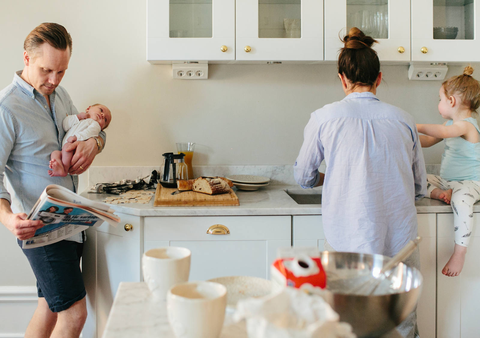 Giovane famiglia è insieme in cucina. Il padre tiene in braccio un neonato e legge il giornale.