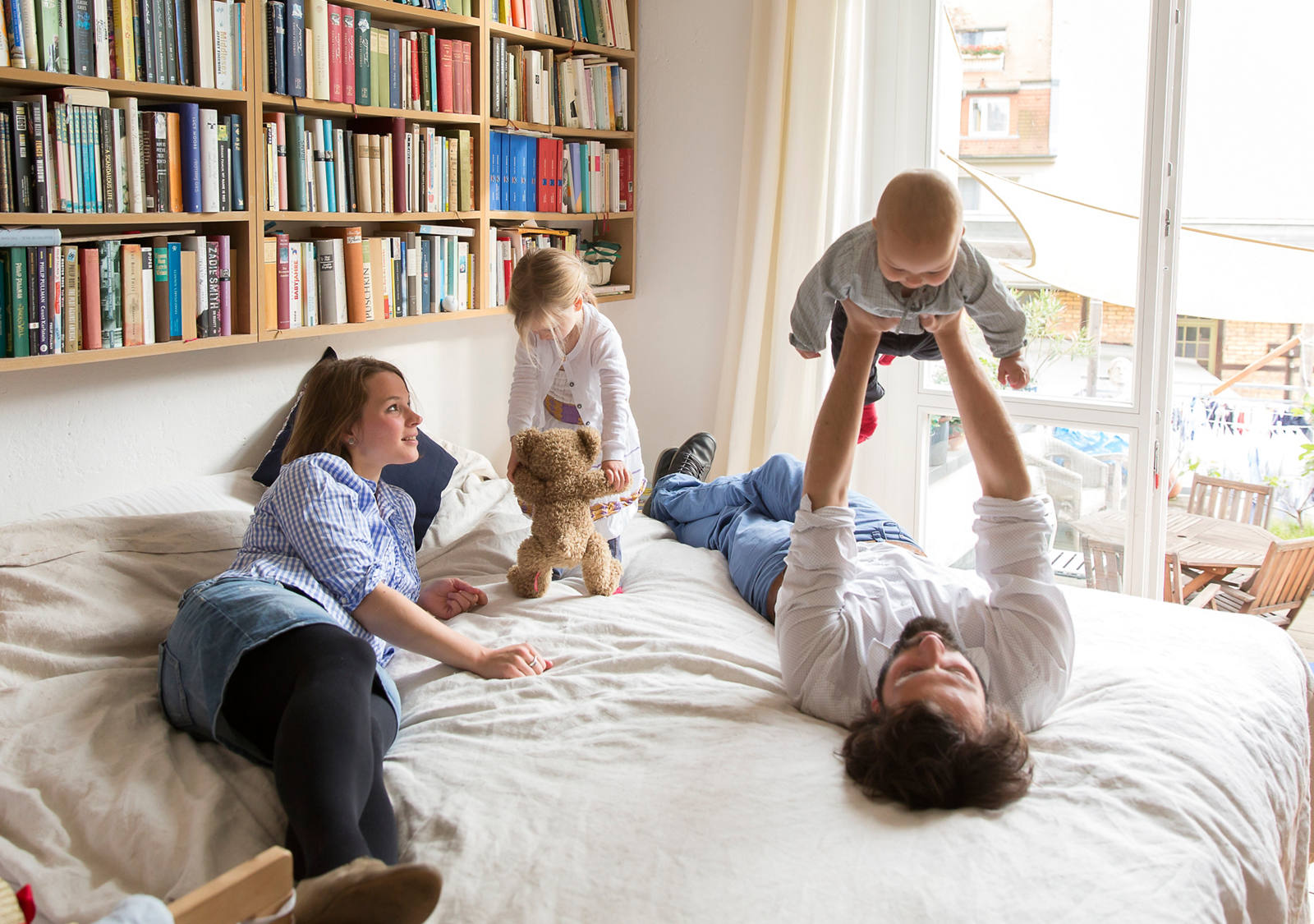 Une jeune famille s’amuse sur le lit des parents. Une bibliothèque est suspendue en arrière-plan.