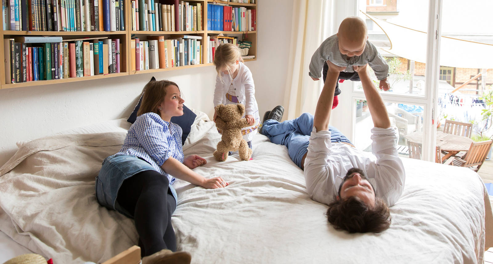 Young family rages on their parents’ bed. A bookshelf hangs in the background.