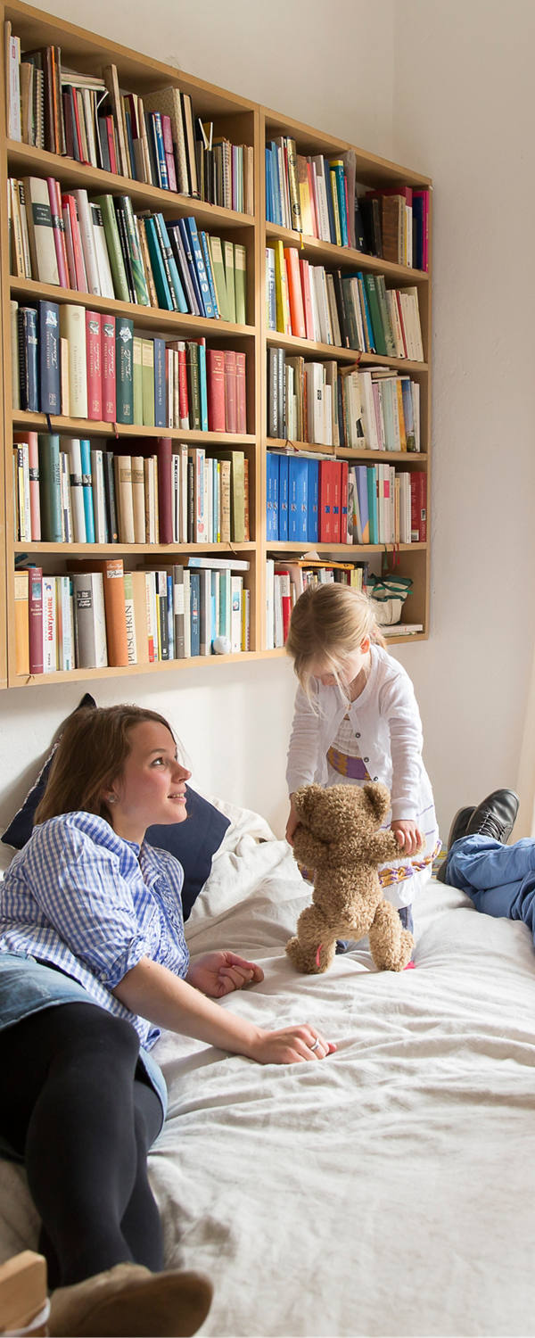 Junge Familie tobt auf dem Elternbett. Im Hintergrund hängt ein Bücherregal.
