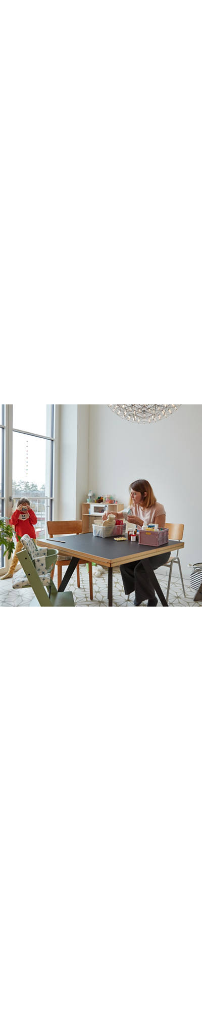 Une femme est assise à la table de la cuisine et range l’armoire à pharmacie. Pendant ce temps, l’enfant joue en pyjama à la fenêtre.