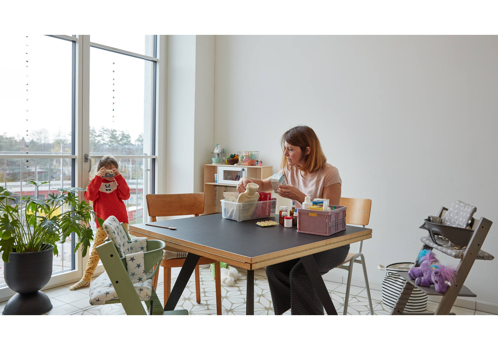 Am Küchentisch sitzt eine Frau und ordnet die Hausapotheke. Währenddessen spielt das Kind im Pyjama am Fenster.