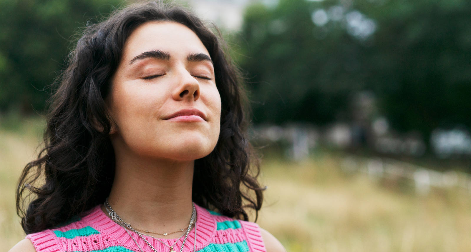 Une femme se tient dans une prairie les yeux fermés et respire profondément.