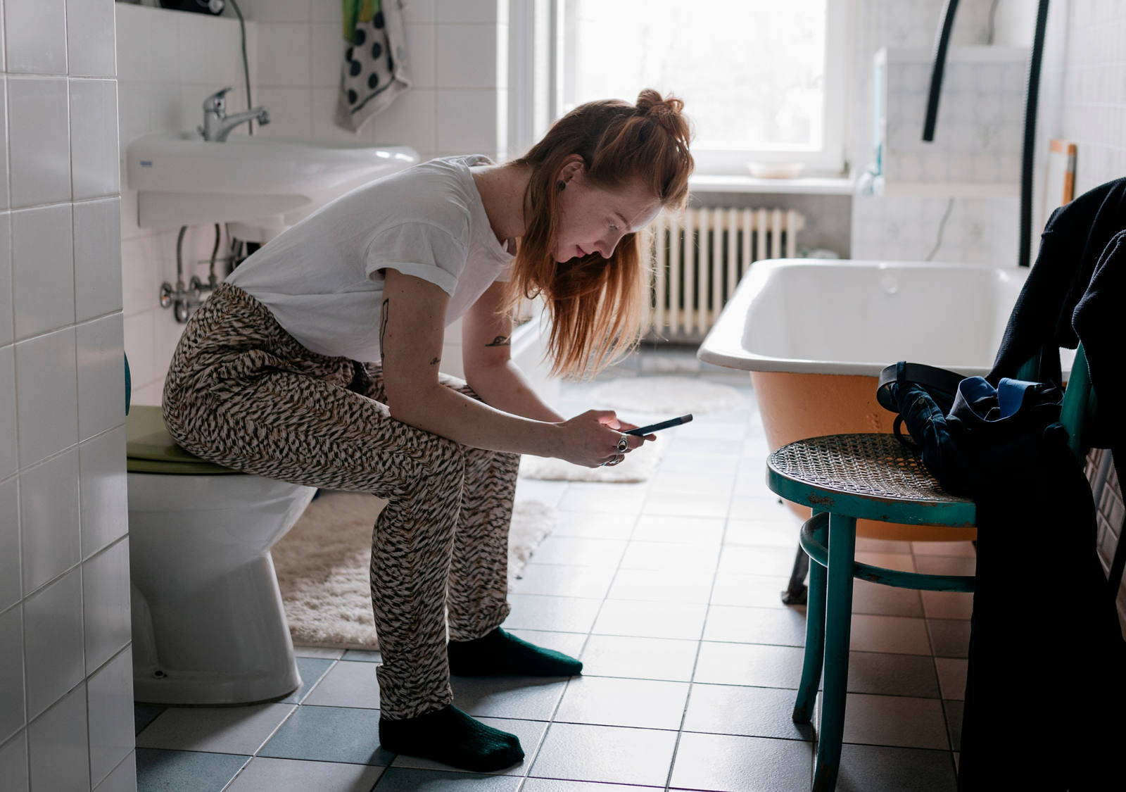 Eine Frau im Badezimmer sitzt auf der Toilette und schaut in ihr Smartphone.