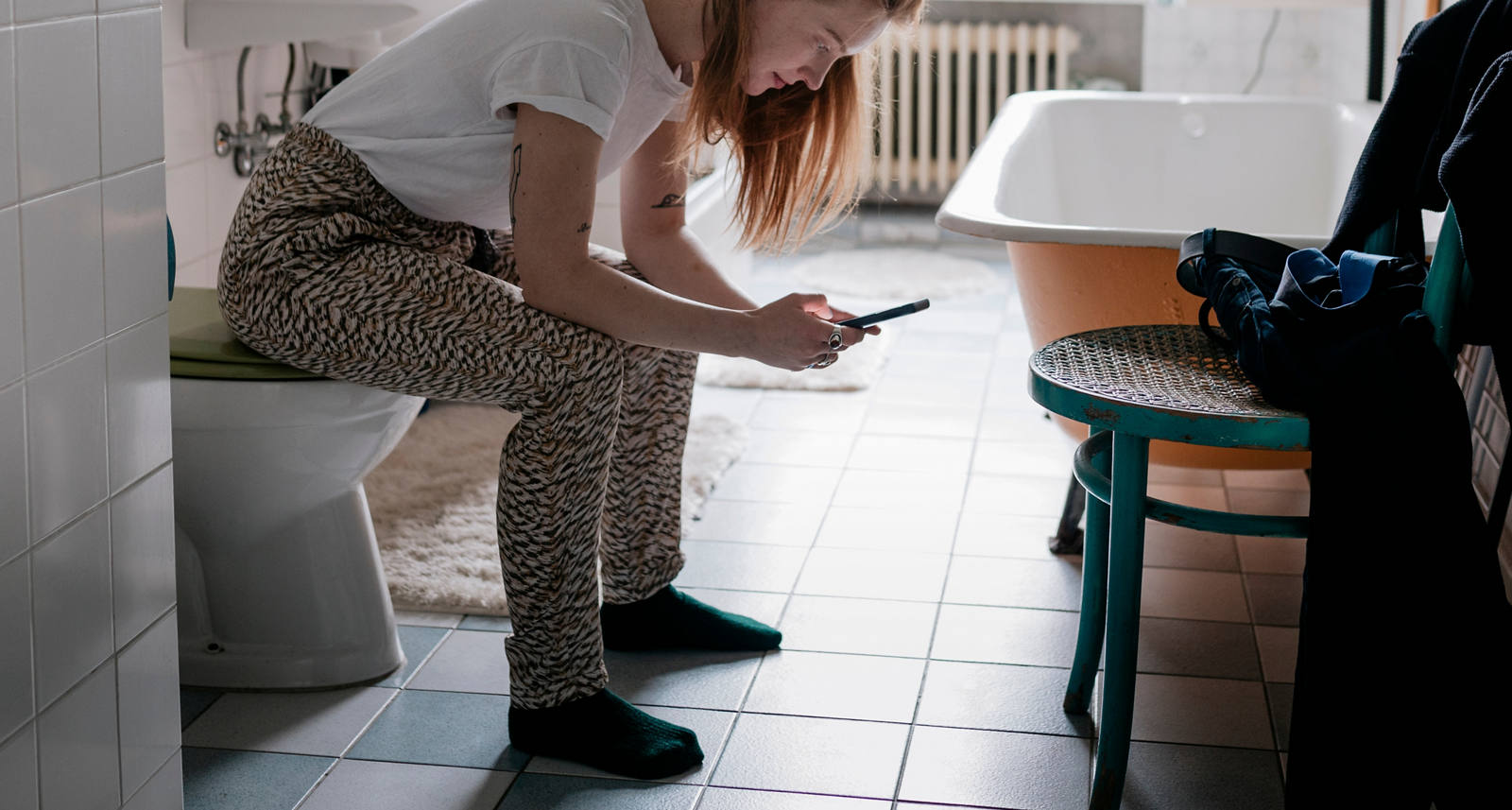 Eine Frau im Badezimmer sitzt auf der Toilette und schaut in ihr Smartphone.