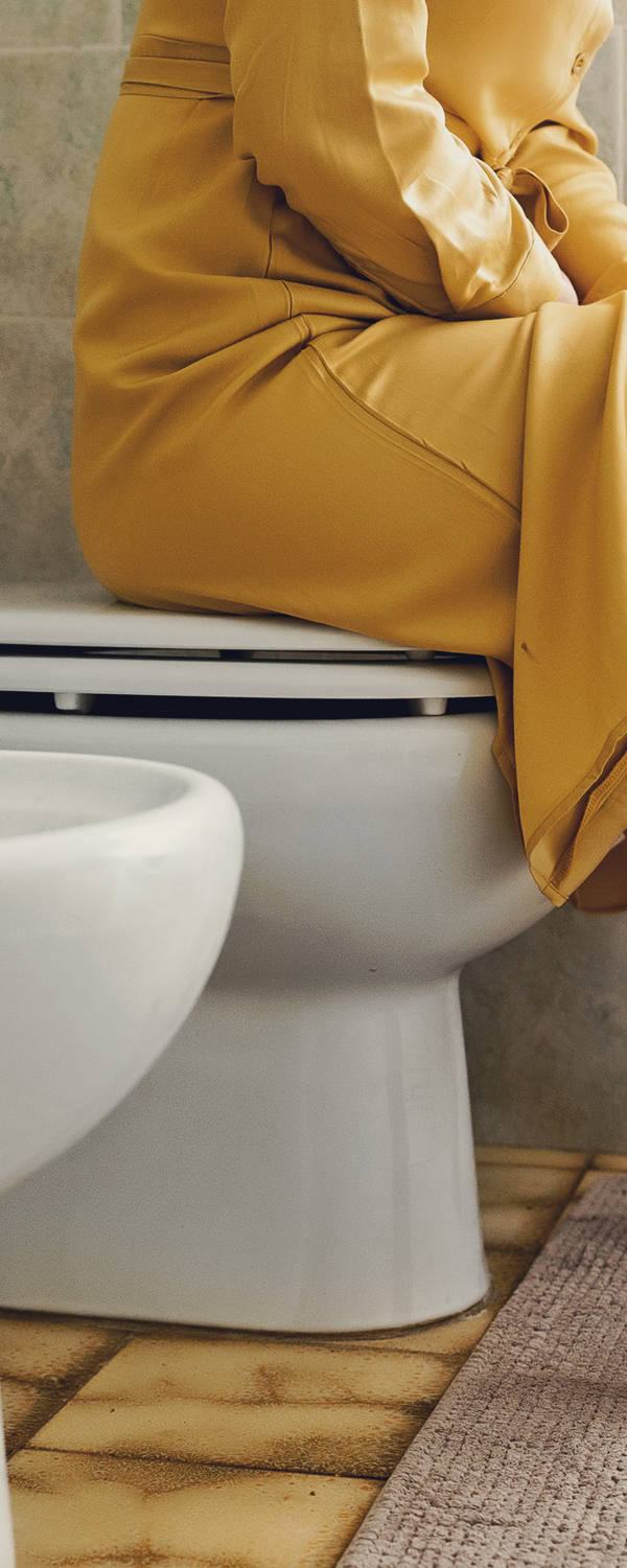 A woman sits on a closed toilet lid in the bathroom.