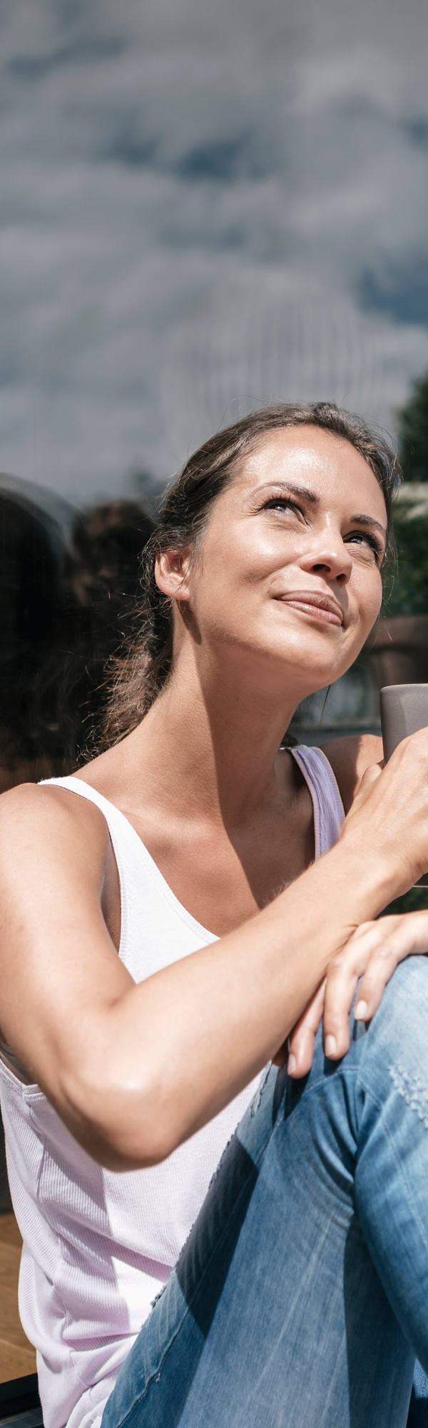 Eine Frau sitzt auf dem Balkon-Boden mit einer Tasse in der Hand und geniesst die Sonnenstrahlen.