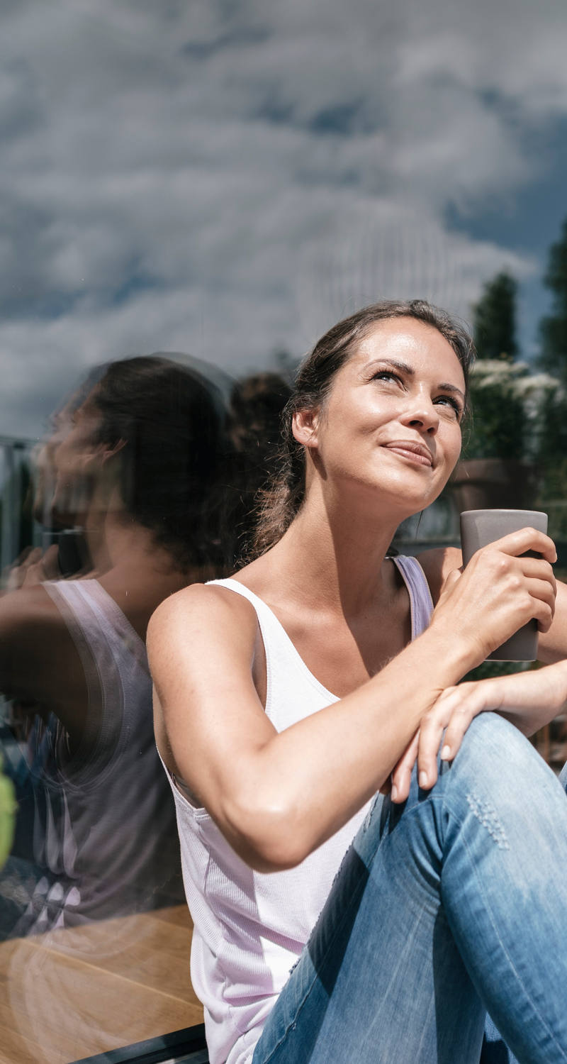 Eine Frau sitzt auf dem Balkon-Boden mit einer Tasse in der Hand und geniesst die Sonnenstrahlen.