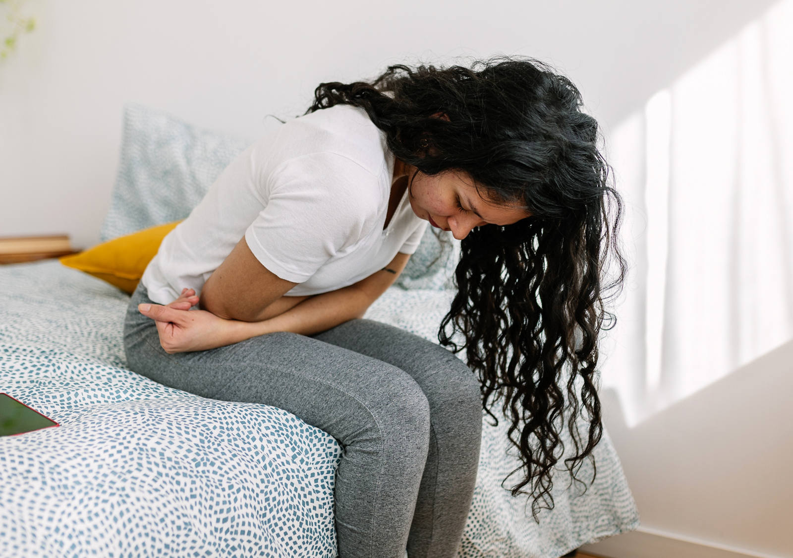 La giovane donna è seduta sul letto, piegata su se stessa, e si tiene la pancia per il dolore.