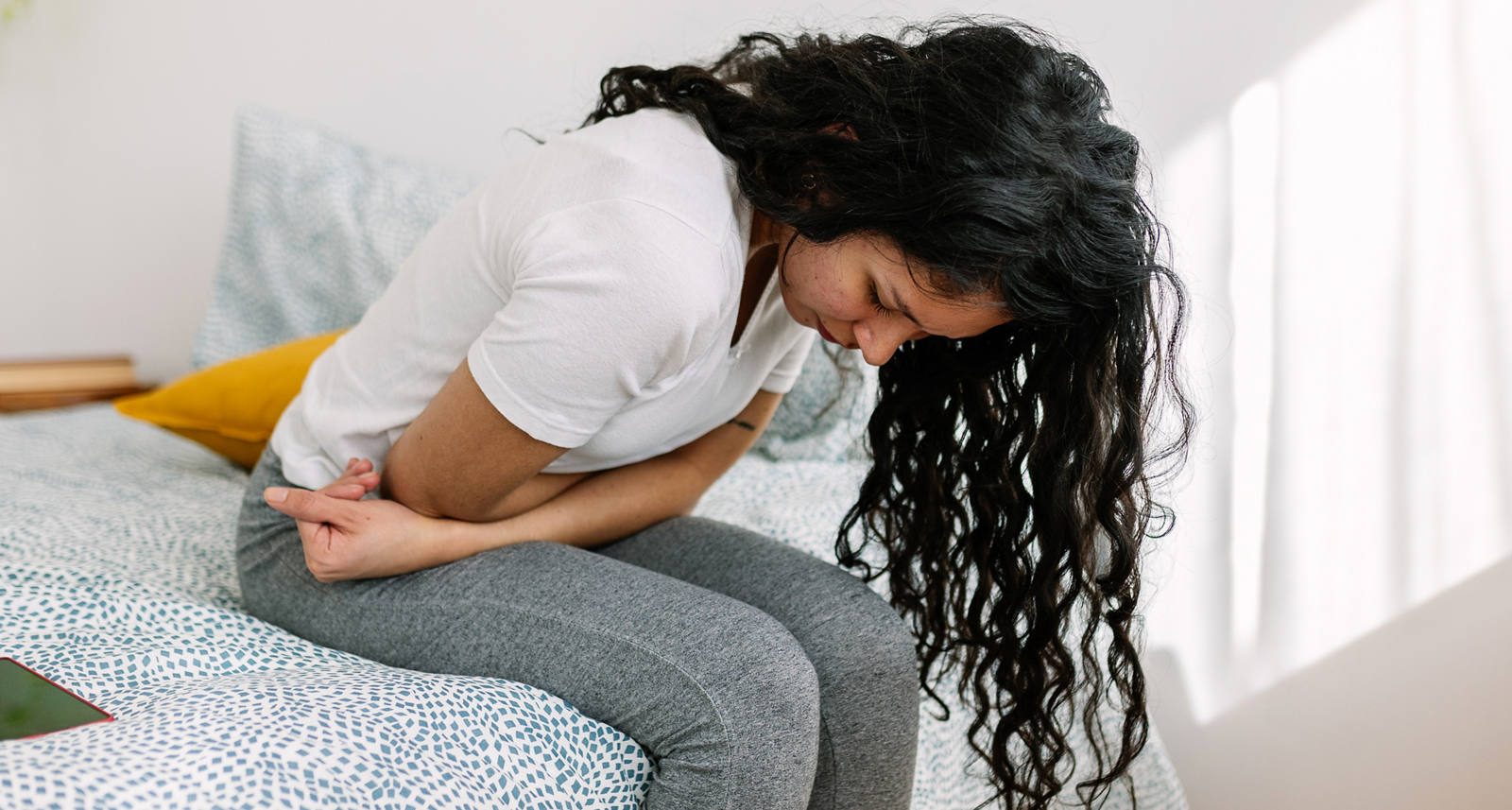 Une jeune femme est assise sur un lit, recroquevillée. Elle se tient le ventre, elle a l’air d’avoir mal.