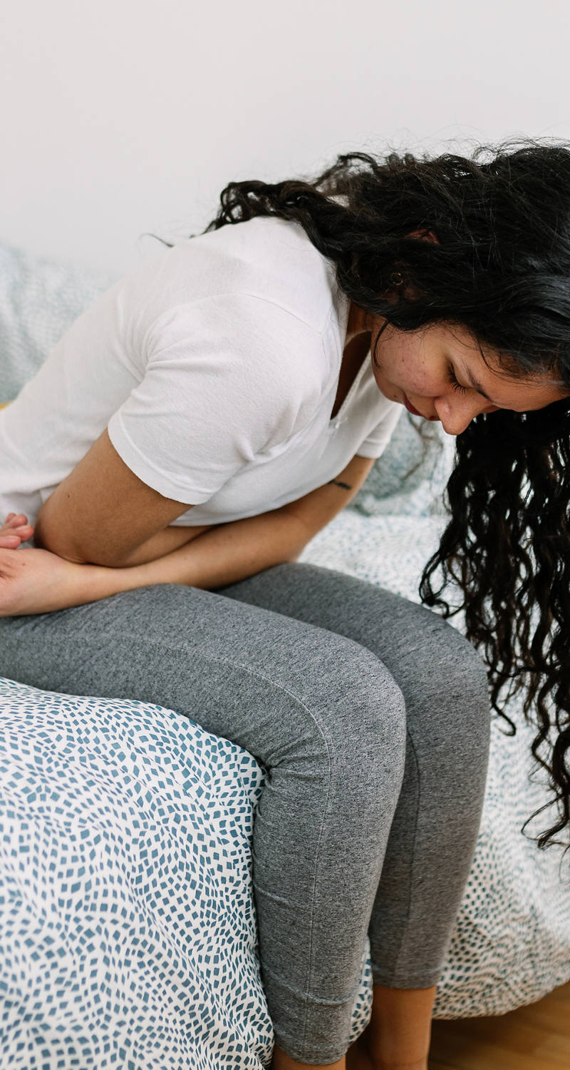 The young woman sits hunched over on the bed, holding her stomach in pain.