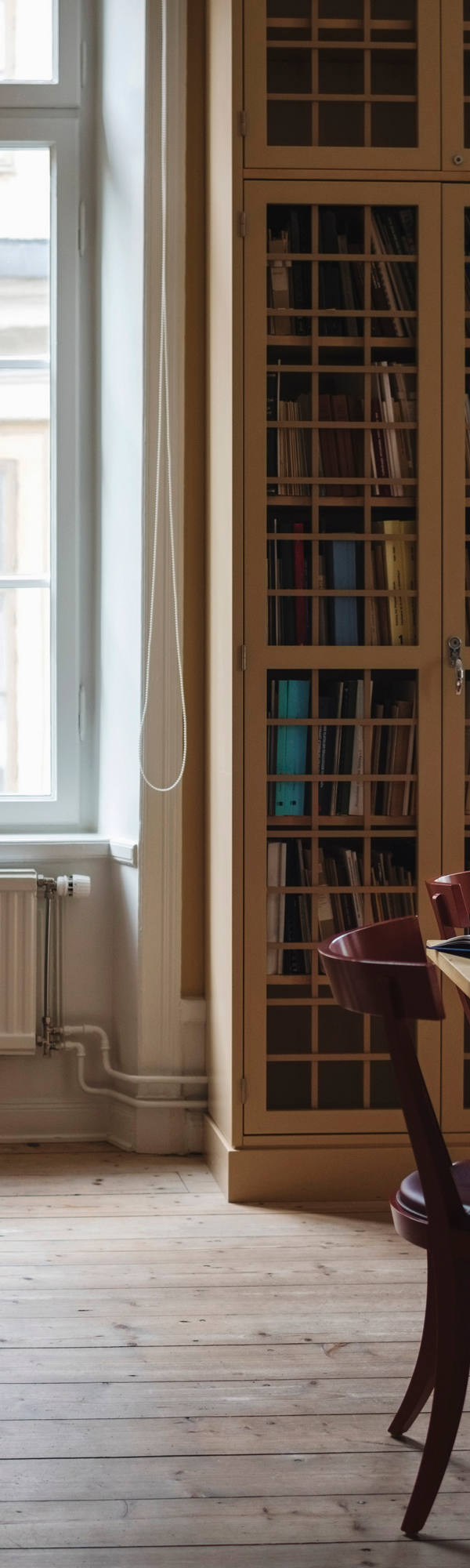 A woman leans backward against a large wall of cupboard, a book in her hand and a pen in her mouth.