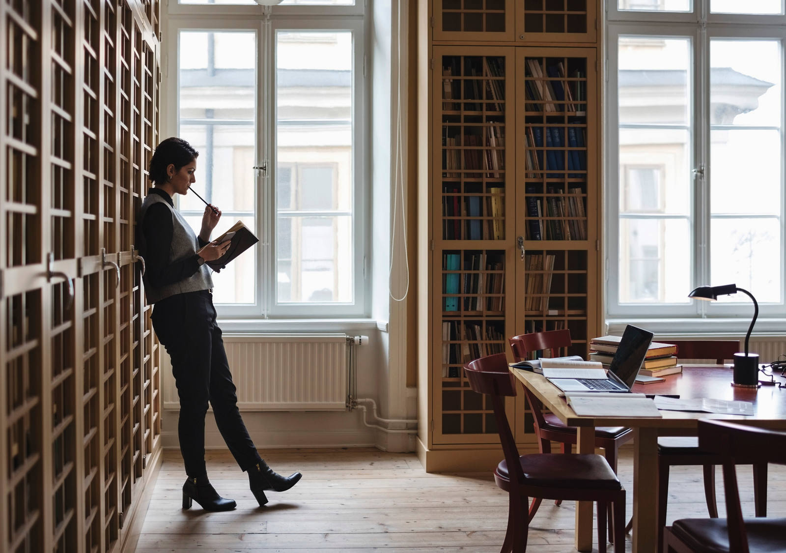 A woman leans backward against a large wall of cupboard, a book in her hand and a pen in her mouth.