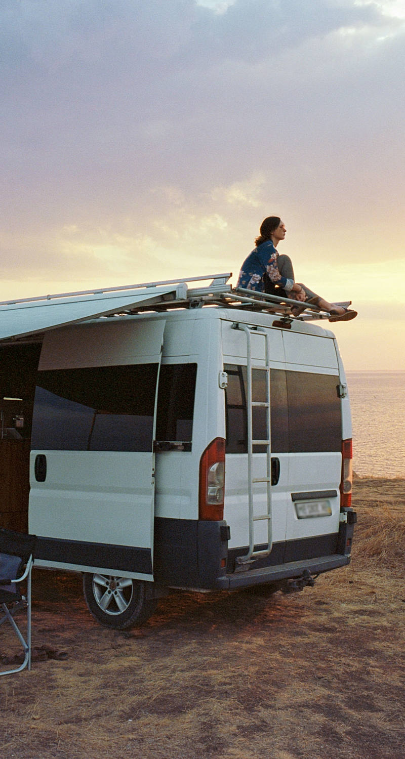 Femme assise sur le toit d’un camping-car et admire le coucher de soleil sur la mer.