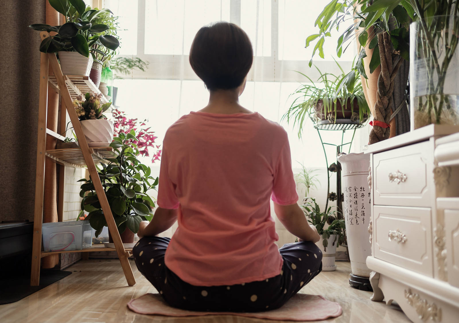 Im Schneidersitz sitzt eine Frau am Boden inmitten einem Raum voller Pflanzen und entspannt sich.