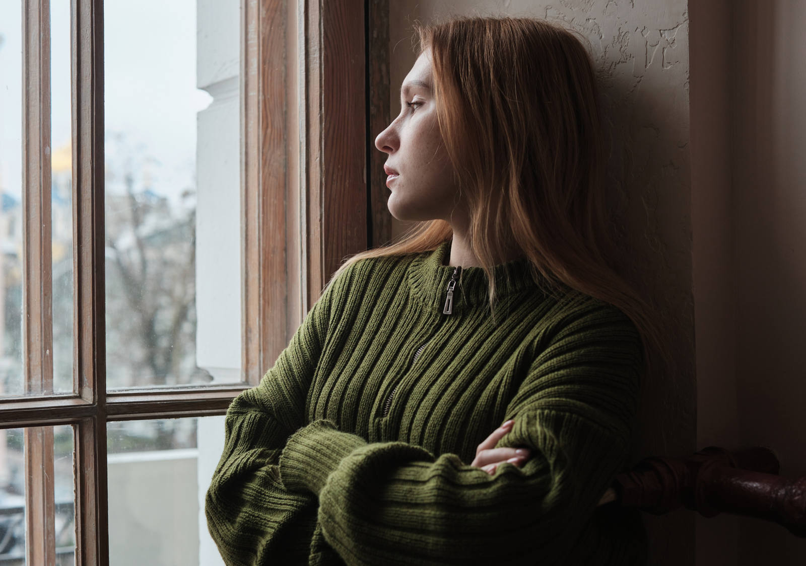 Eine junge Frau lehnt sich mit verschränkten Armen an die Wand und schaut nachdenklich aus dem Fenster.