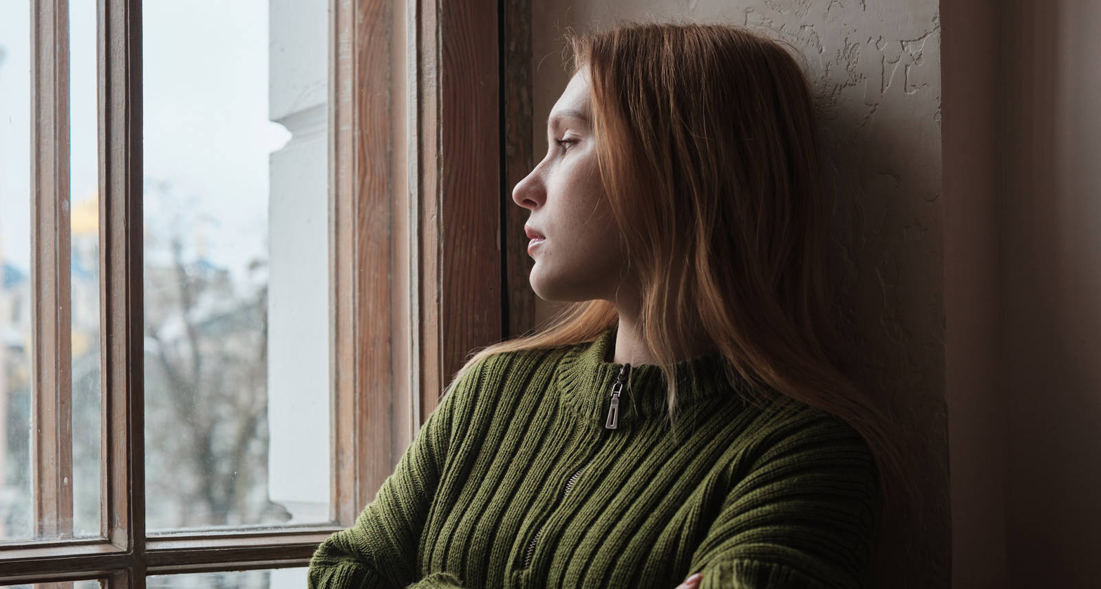 Eine junge Frau lehnt sich mit verschränkten Armen an die Wand und schaut nachdenklich aus dem Fenster.