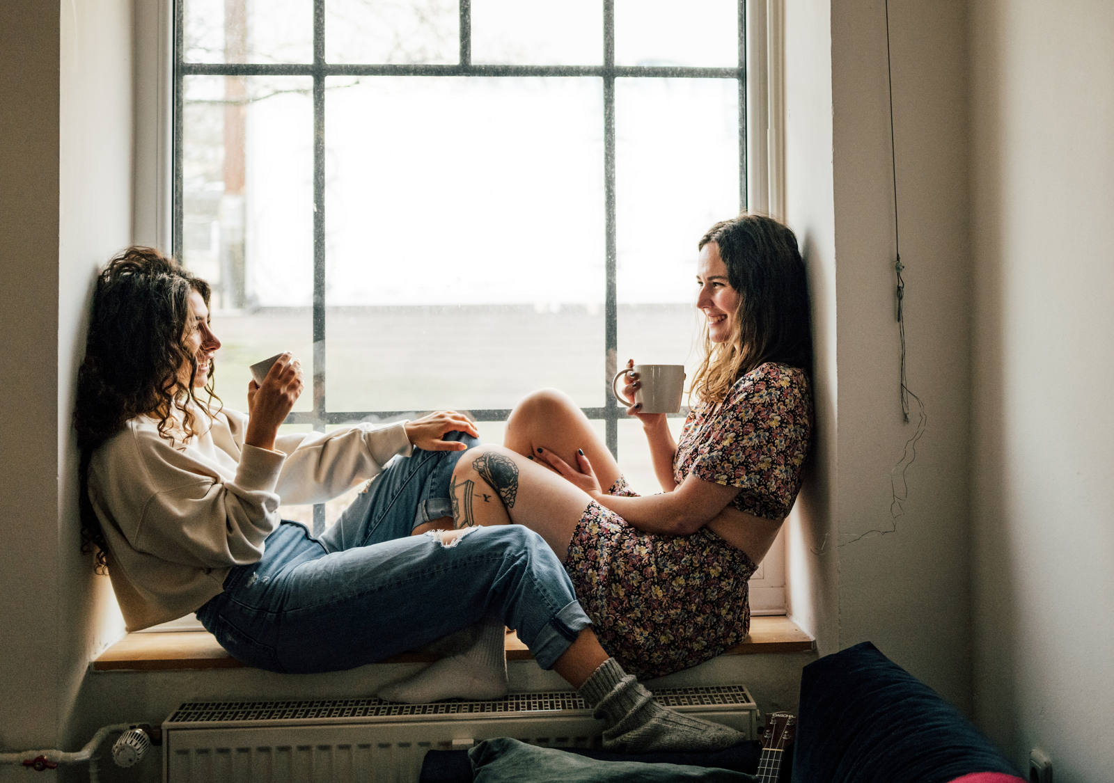 Zwei Frauen sitzen auf Fenstersims, trinken ein warmes Getränk und geniessen die Zeit zusammen.