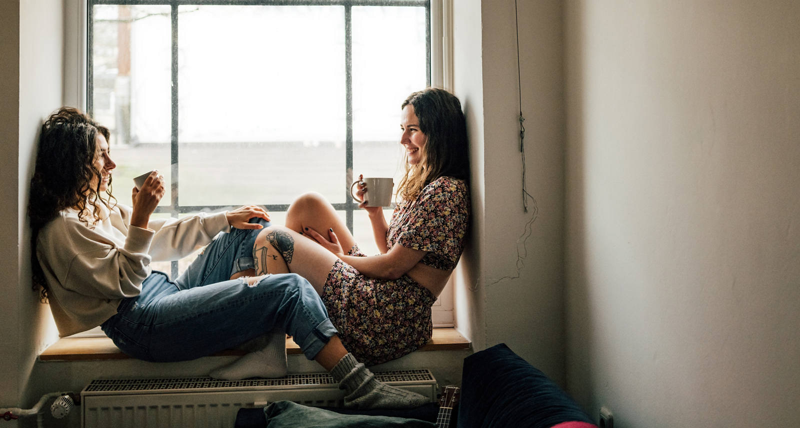 Zwei Frauen sitzen auf Fenstersims, trinken ein warmes Getränk und geniessen die Zeit zusammen.