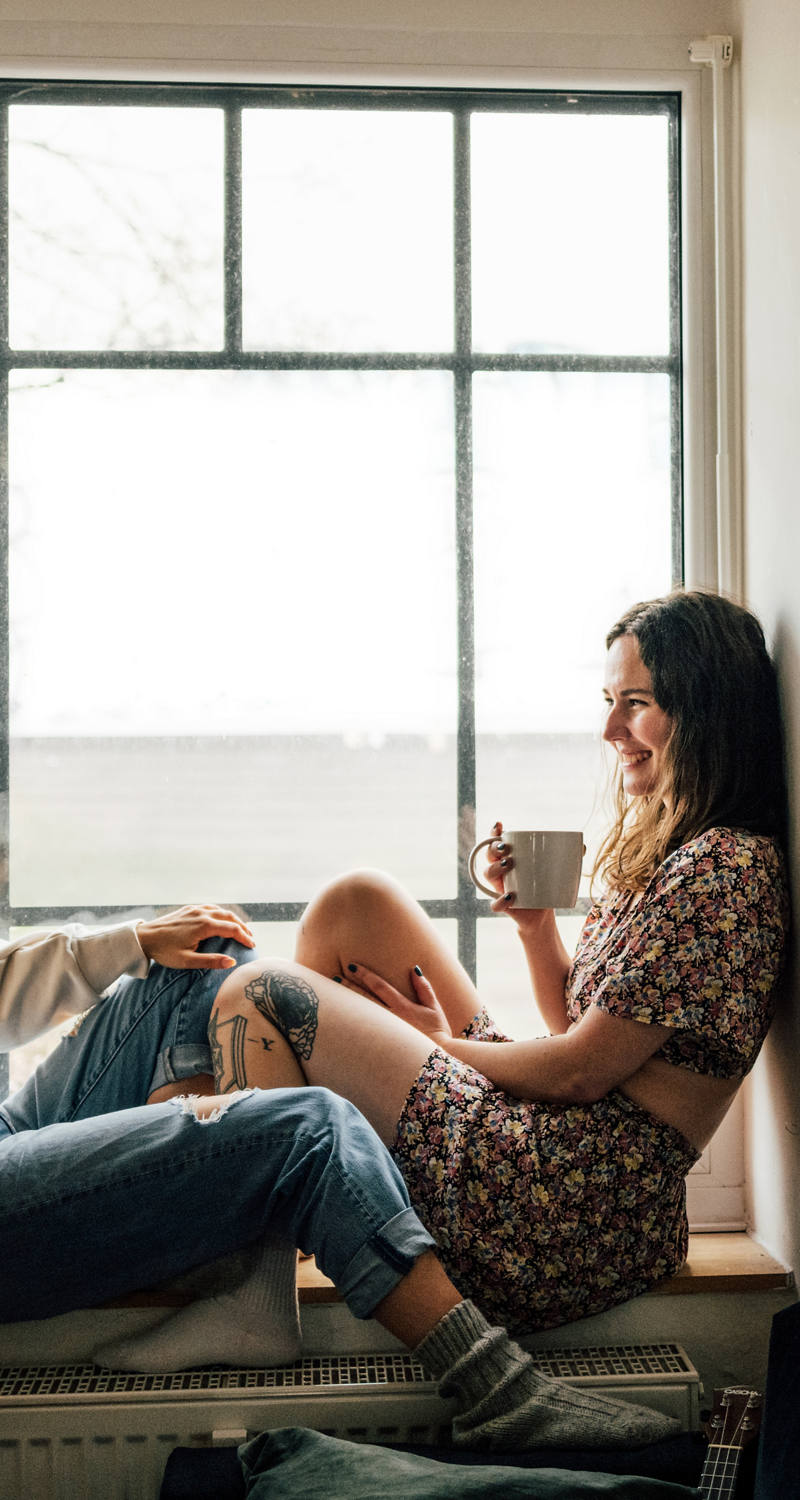 Zwei Frauen sitzen auf Fenstersims, trinken ein warmes Getränk und geniessen die Zeit zusammen.