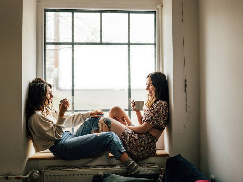 Zwei Frauen sitzen auf Fenstersims, trinken ein warmes Getränk und geniessen die Zeit zusammen.
