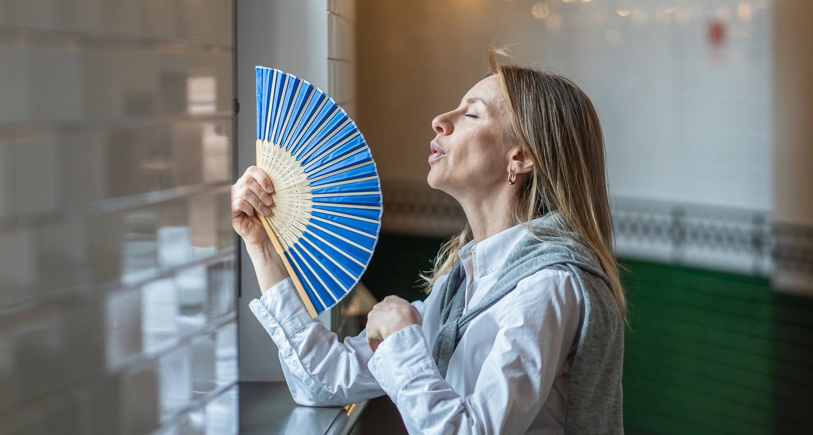 Frau steht an einem heissen Tag im Flur und versucht mit ihrem Handfächer ihre hohe Körpertemperatur zu lindern, damit sie nicht so stark schwitzt.