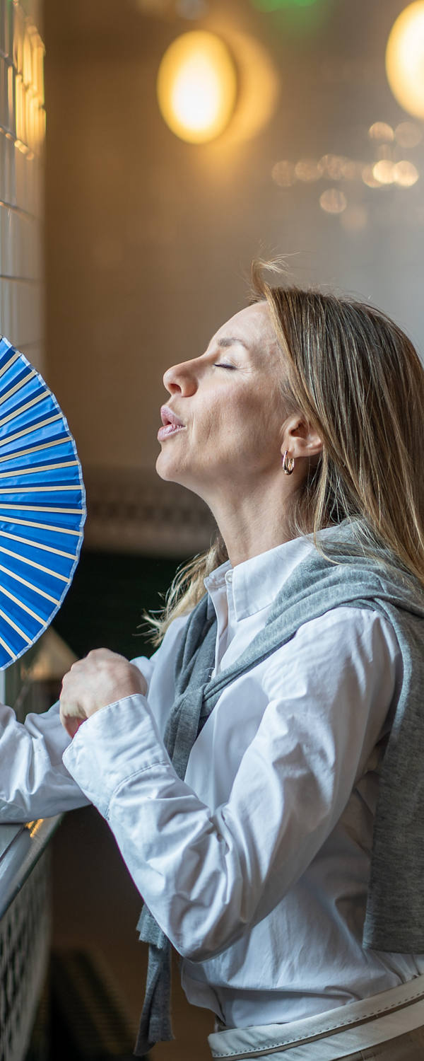 Frau steht an einem heissen Tag im Flur und versucht mit ihrem Handfächer ihre hohe Körpertemperatur zu lindern, damit sie nicht so stark schwitzt.