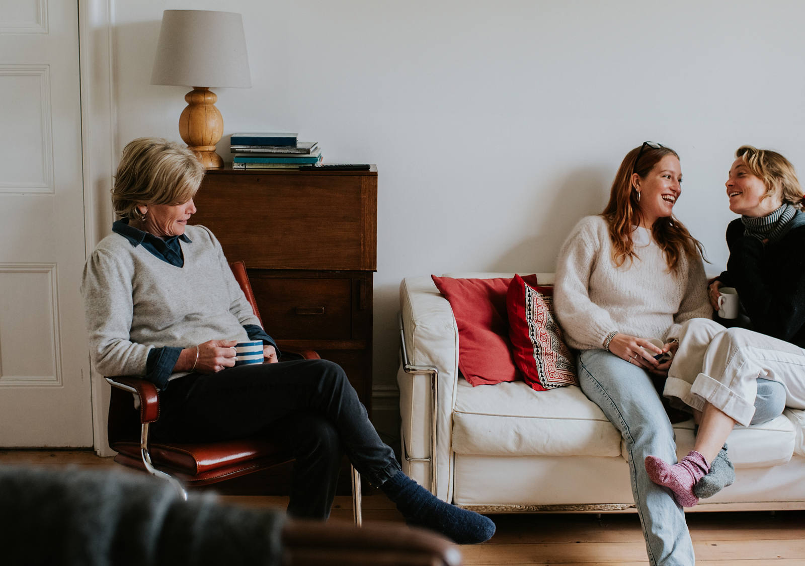 Two young women are sitting on the sofa in the living room, laughing with each other. An older woman nearby is watching them, holding a cup on her lap.