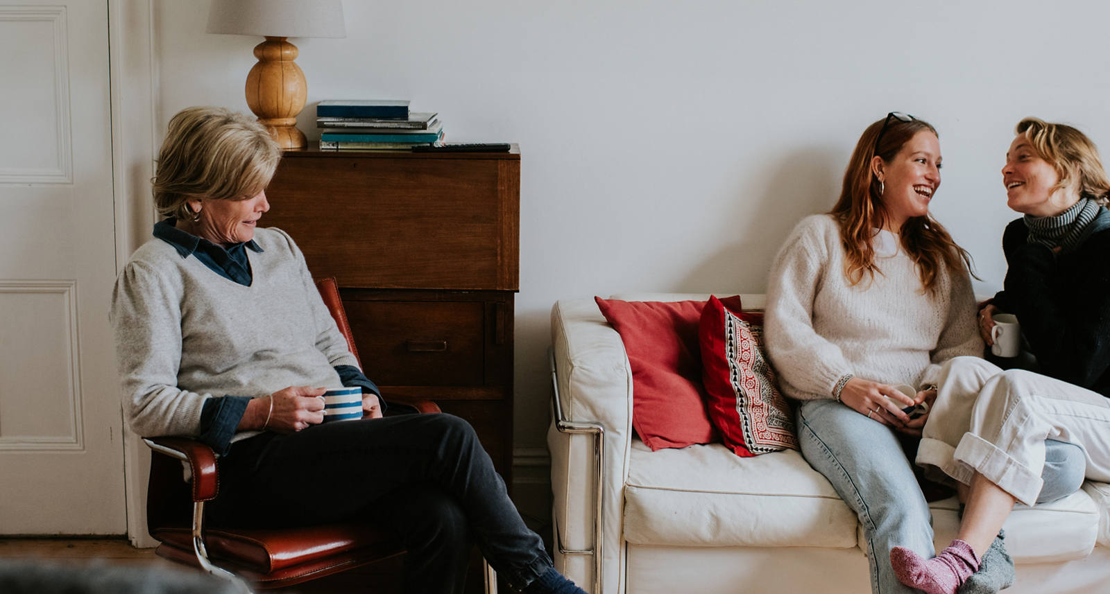 Two young women are sitting on the sofa in the living room, laughing with each other. An older woman nearby is watching them, holding a cup on her lap.