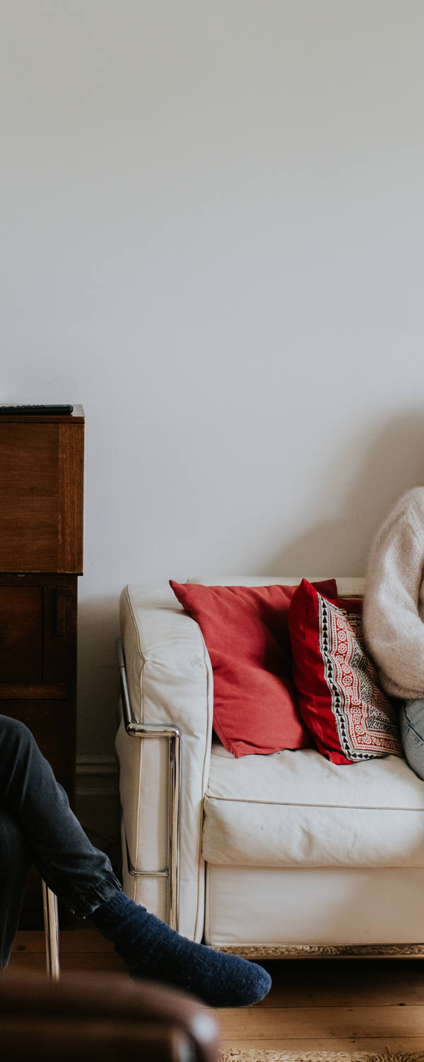 Zwei junge Frauen sitzen im Wohnzimmer auf dem Sofa und lachen miteinander. Eine ältere Dame daneben schaut die beiden anderen an und hält eine Tasse auf ihrem Schoss.