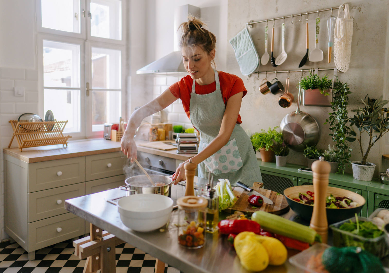 Eine Frau mit Kochschürze steht in der Küche und bereitet eine Mahlzeit mit viel Gemüse zu.
