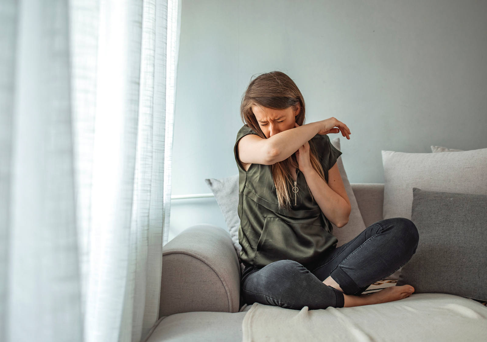 Una donna ammalata è seduta sul divano e con il braccio piega del gomito copre naso e bocca.