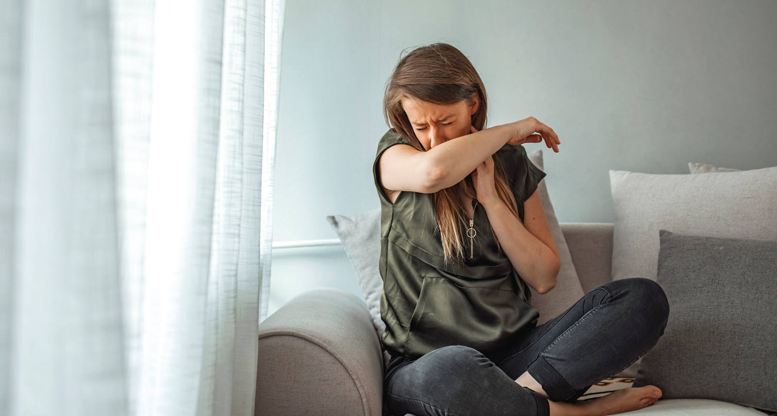 Eine kranke Frau sitzt auf dem Sofa und hält schützend den Ellenbogen vor ihr Gesicht, während sie hustet.