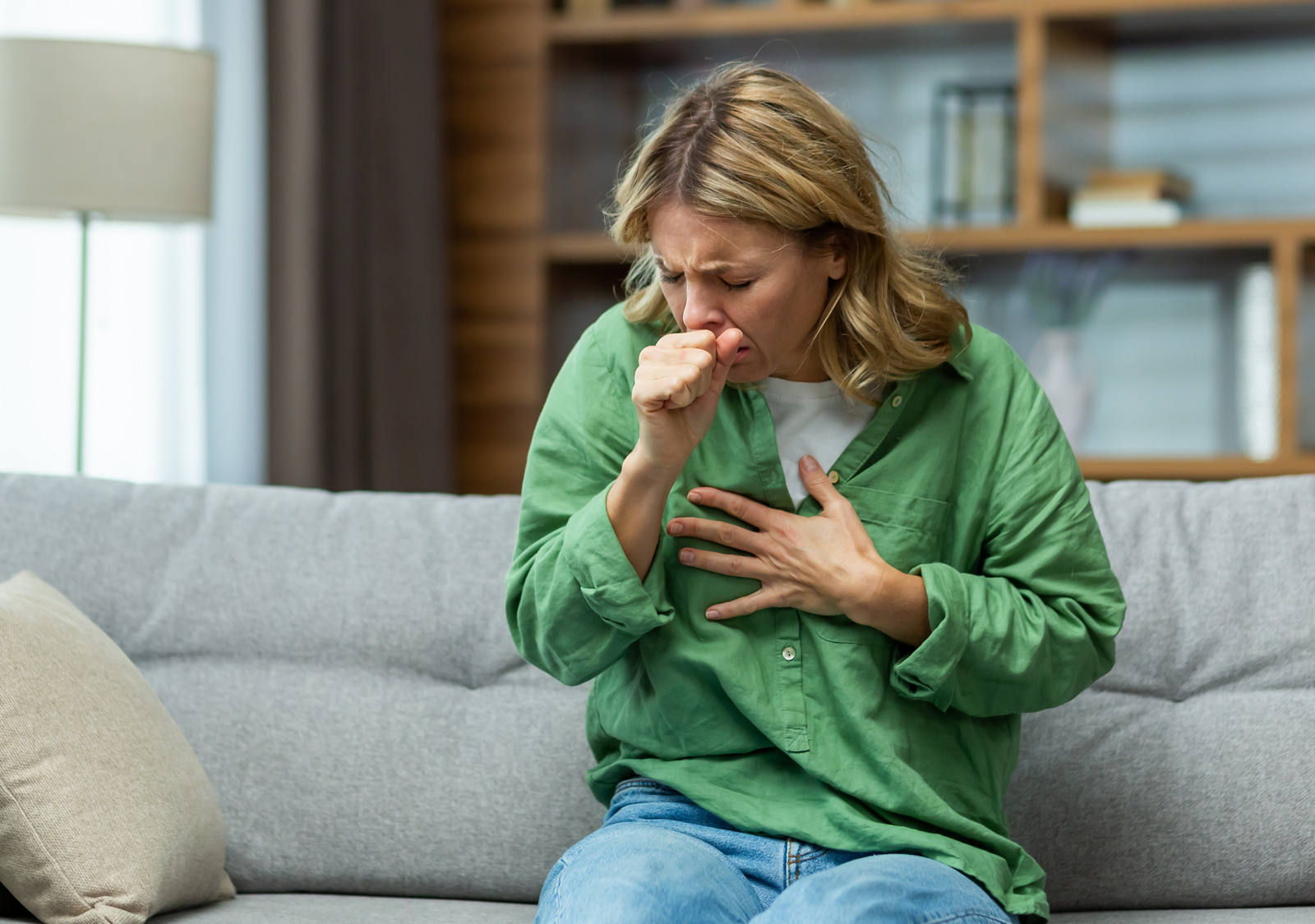 A woman is sitting on the sofa, coughing hard while holding one hand in front of her chest.