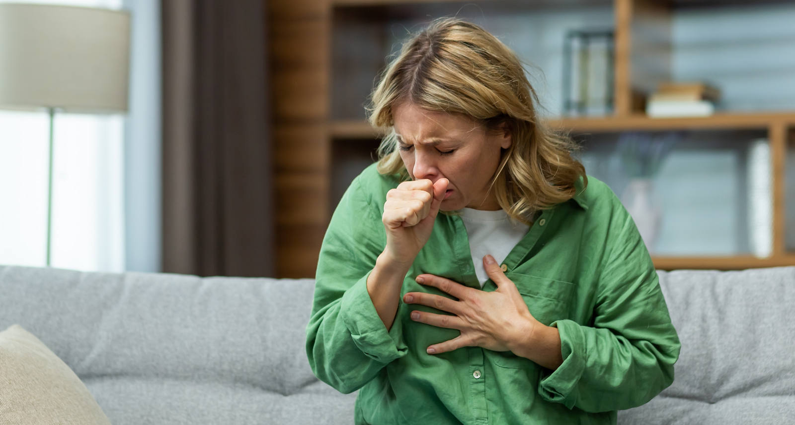 A woman is sitting on the sofa, coughing hard while holding one hand in front of her chest.