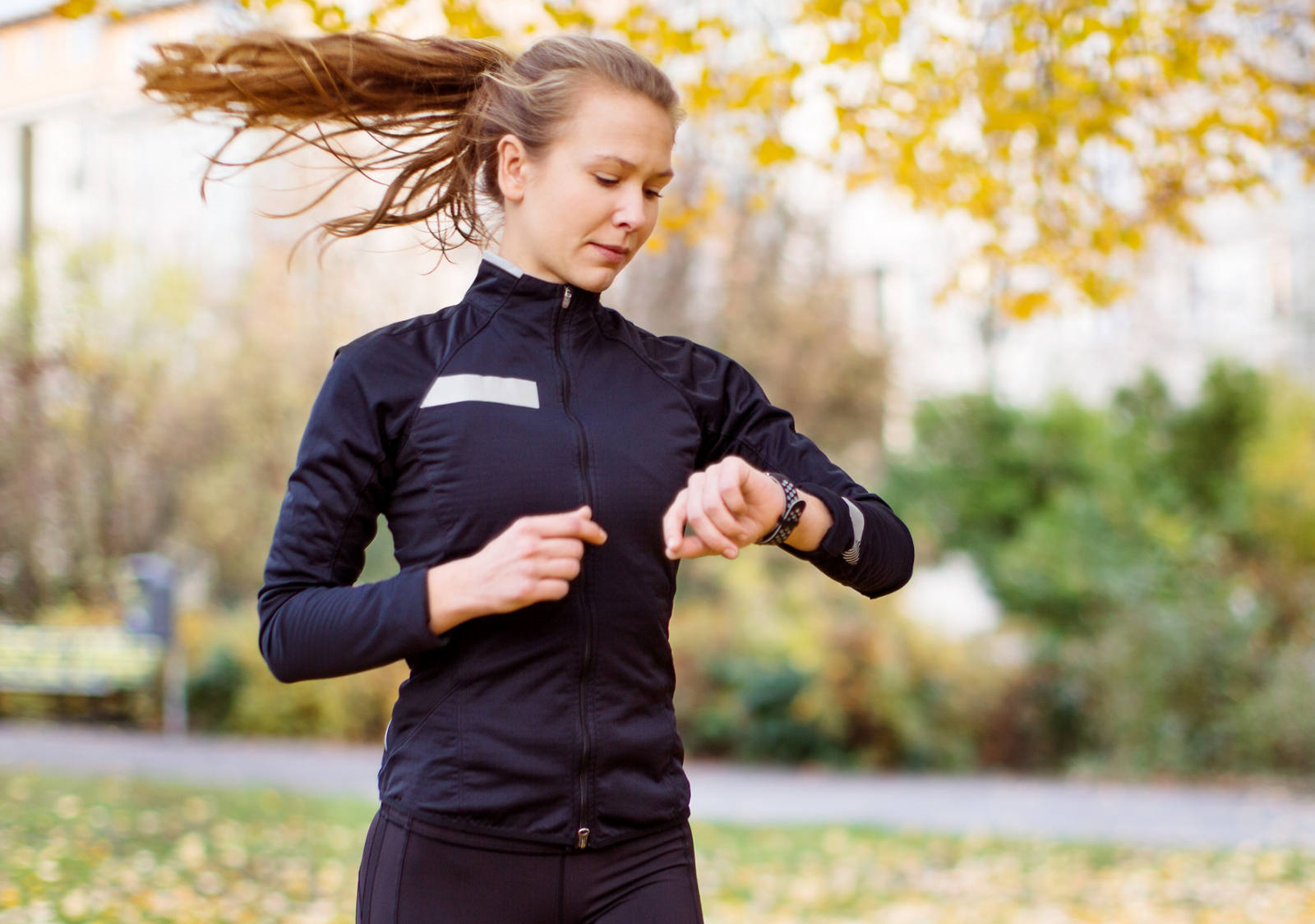 Donna che fa jogging corre sul prato con le foglie autunnali e controlla il suo smartwatch.