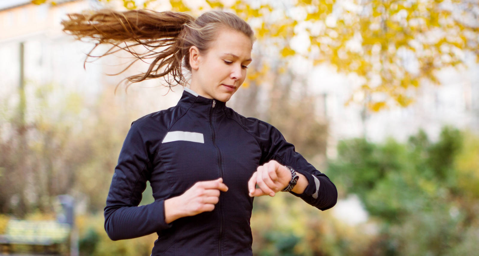 Une coureuse marche dans la prairie avec des feuilles d’automne sur le sol et regarde sa montre connectée.