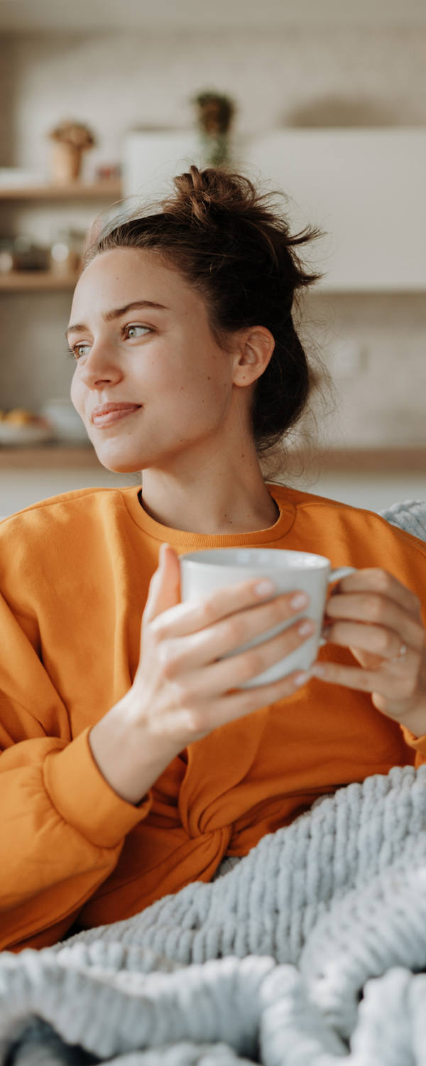 A young woman is sitting on the sofa wrapped in a soft blanket and drinking tea.