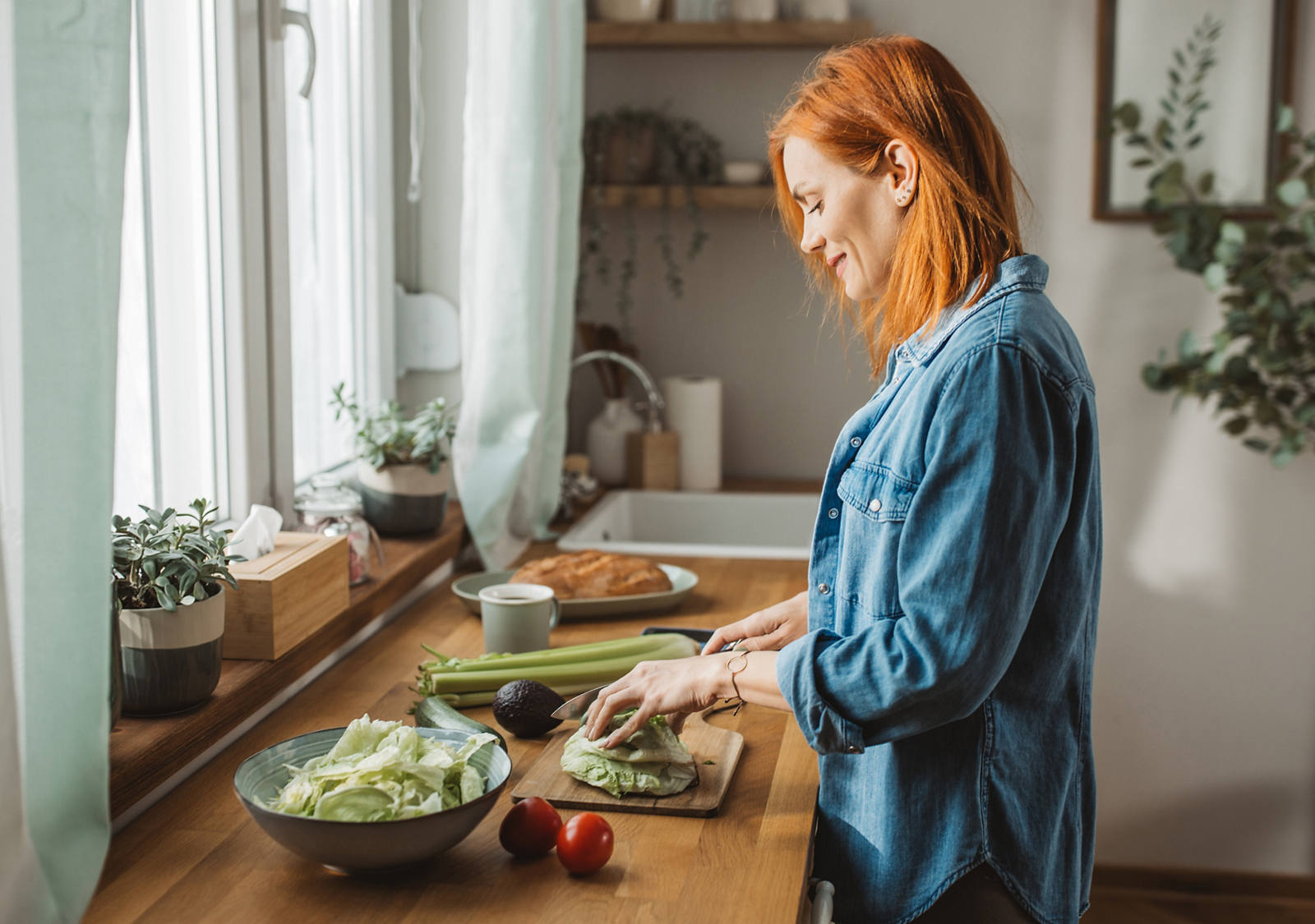Une femme se tient dans la cuisine au soleil et coupe des légumes pour une salade.