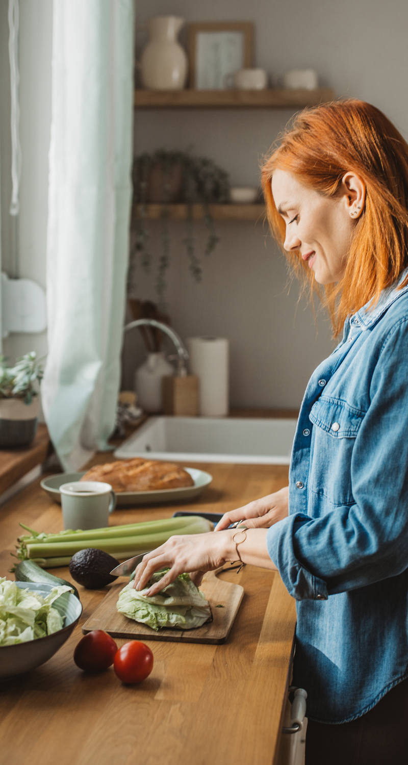 Une femme se tient dans la cuisine au soleil et coupe des légumes pour une salade.