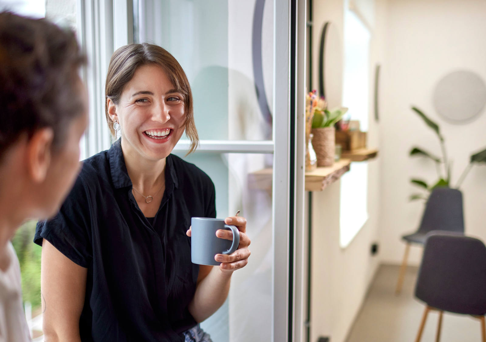Frau lacht im Gespräch mit einem Mann am Fenster und hält einen Becher in der Hand