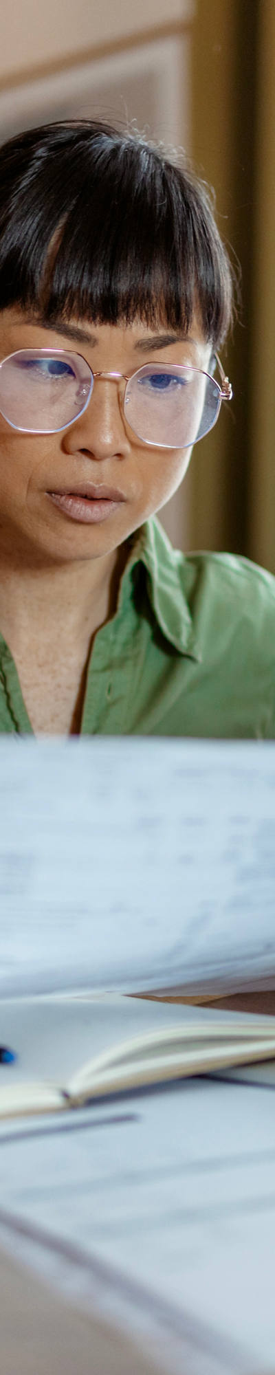 A woman of Asian descent with glasses sits at a round coffee table working with paper and laptop.