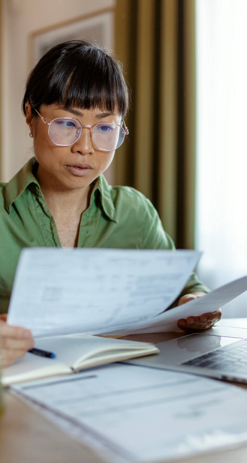 A woman of Asian descent with glasses sits at a round coffee table working with paper and laptop.