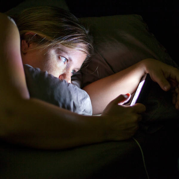 Eine junge Frau liegt im Bett und hat ihr Blick auf das leuchtende Smarphone gerichtet.