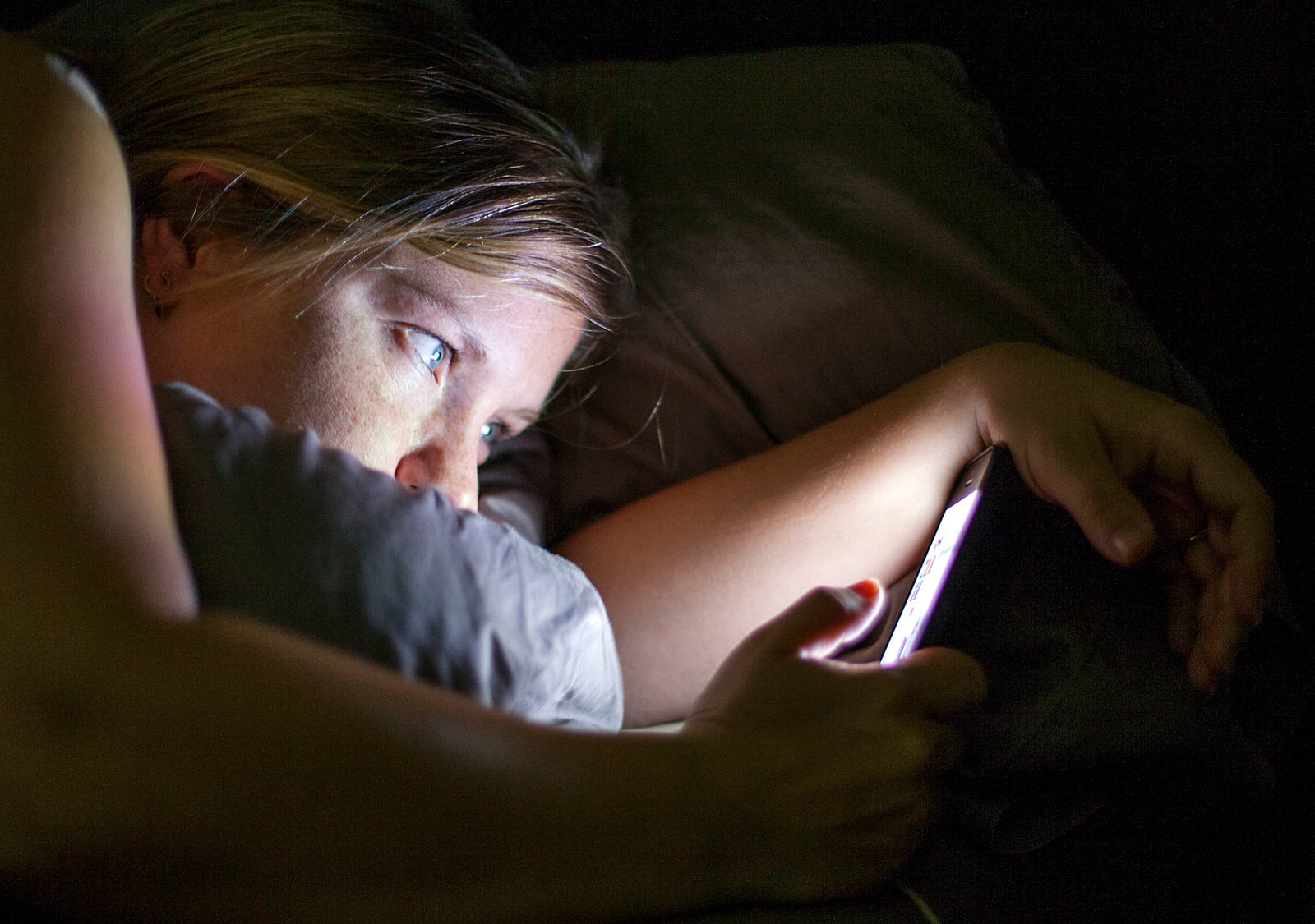 Eine junge Frau liegt im Bett und hat ihr Blick auf das leuchtende Smarphone gerichtet.