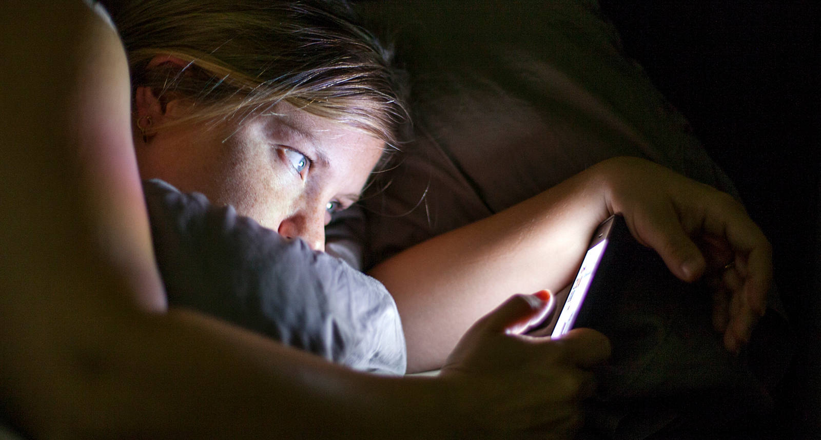 Eine junge Frau liegt im Bett und hat ihr Blick auf das leuchtende Smarphone gerichtet.
